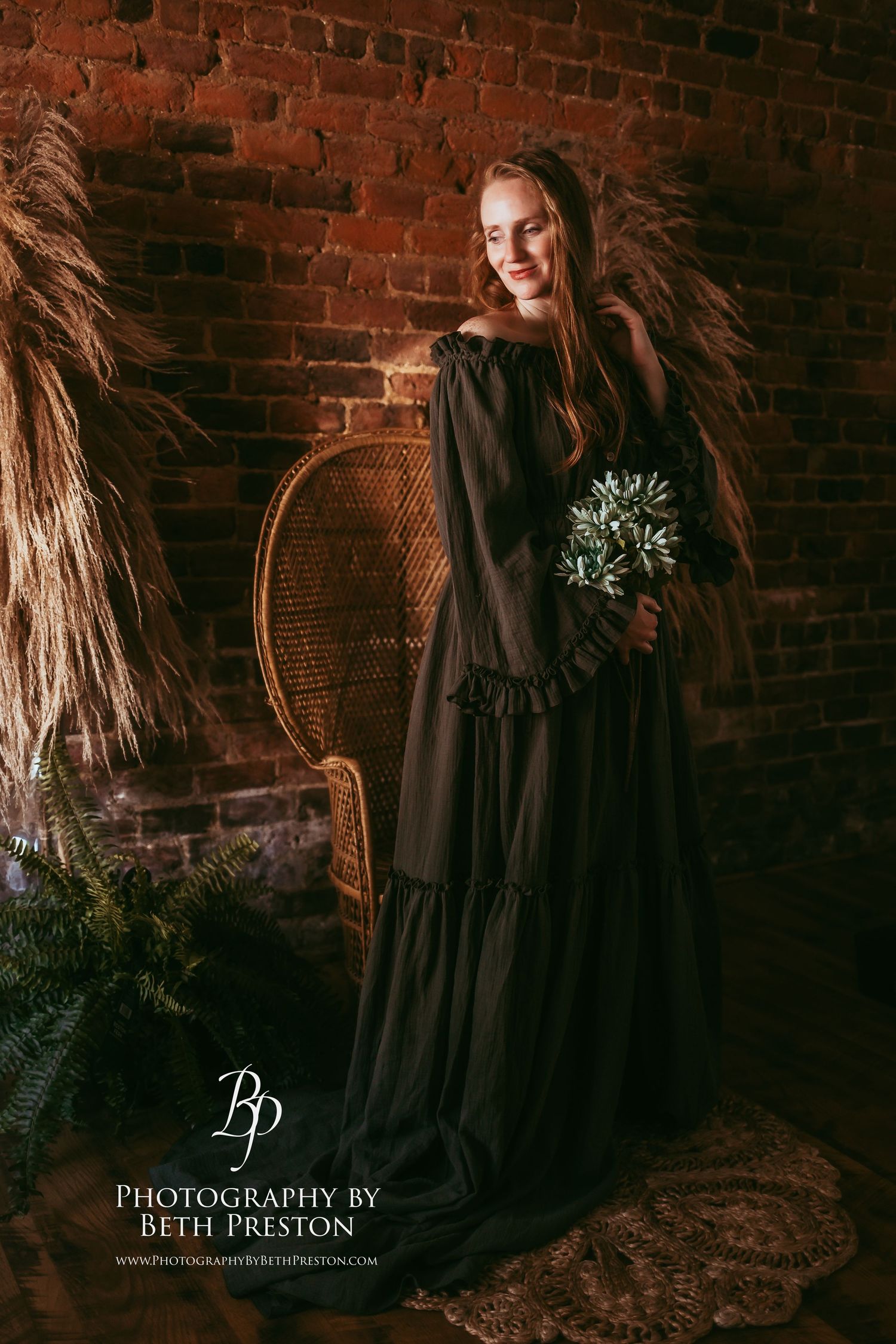 A woman in a flowing black dress holds greenery against a rustic brick wall setting.