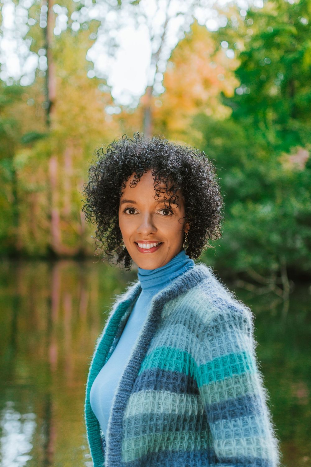 Portrait photograph of a woman with curly hair smiling over her shoulder in front of a lake in the forest.