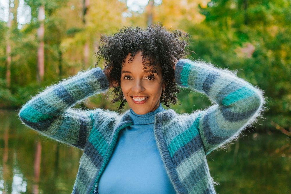 Portrait photograph of a woman smiling while playing with her curly hair in front of a lake in the forest.