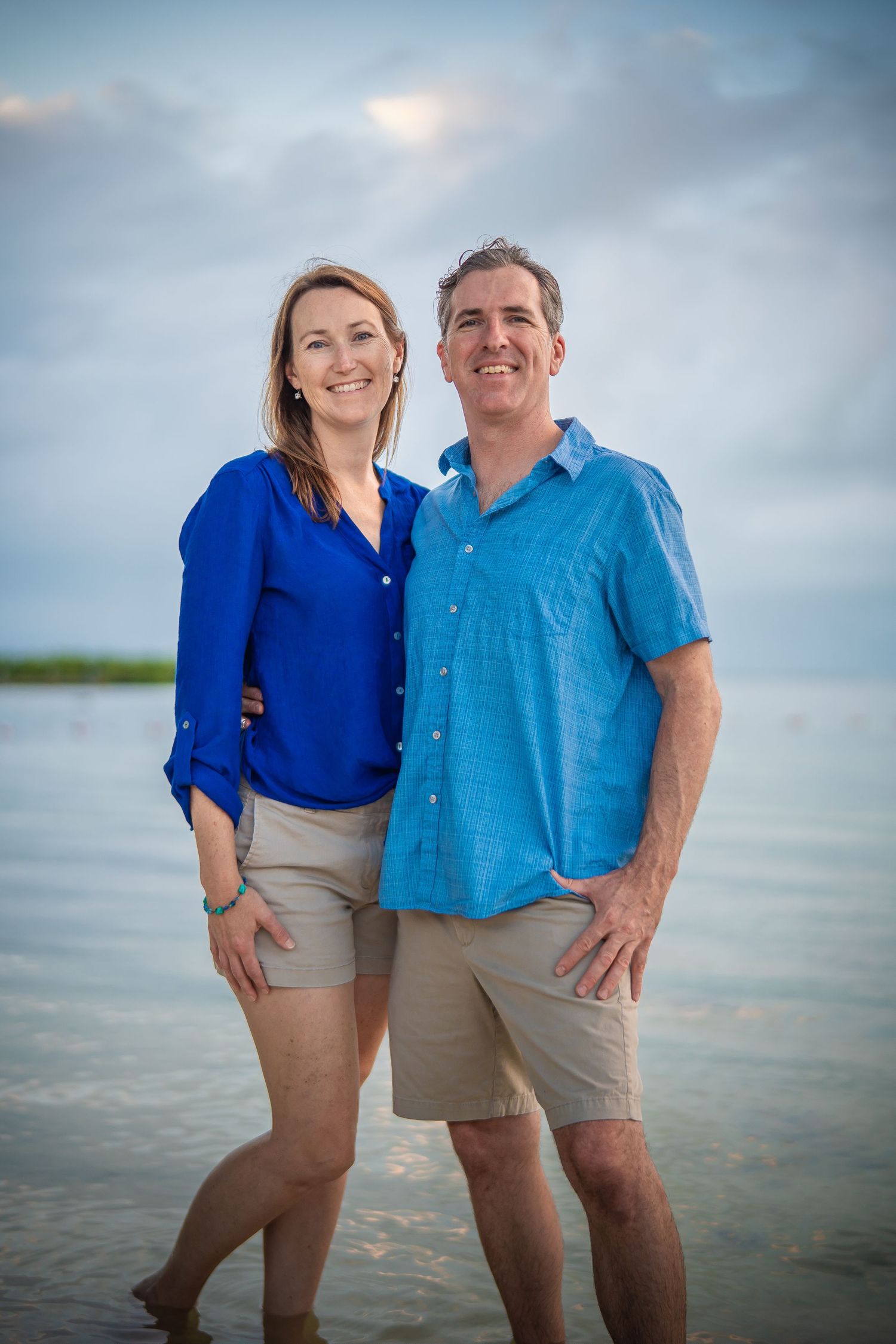 Couple poses together at sunset on a beach in coordinating blue shirts and khaki shorts.