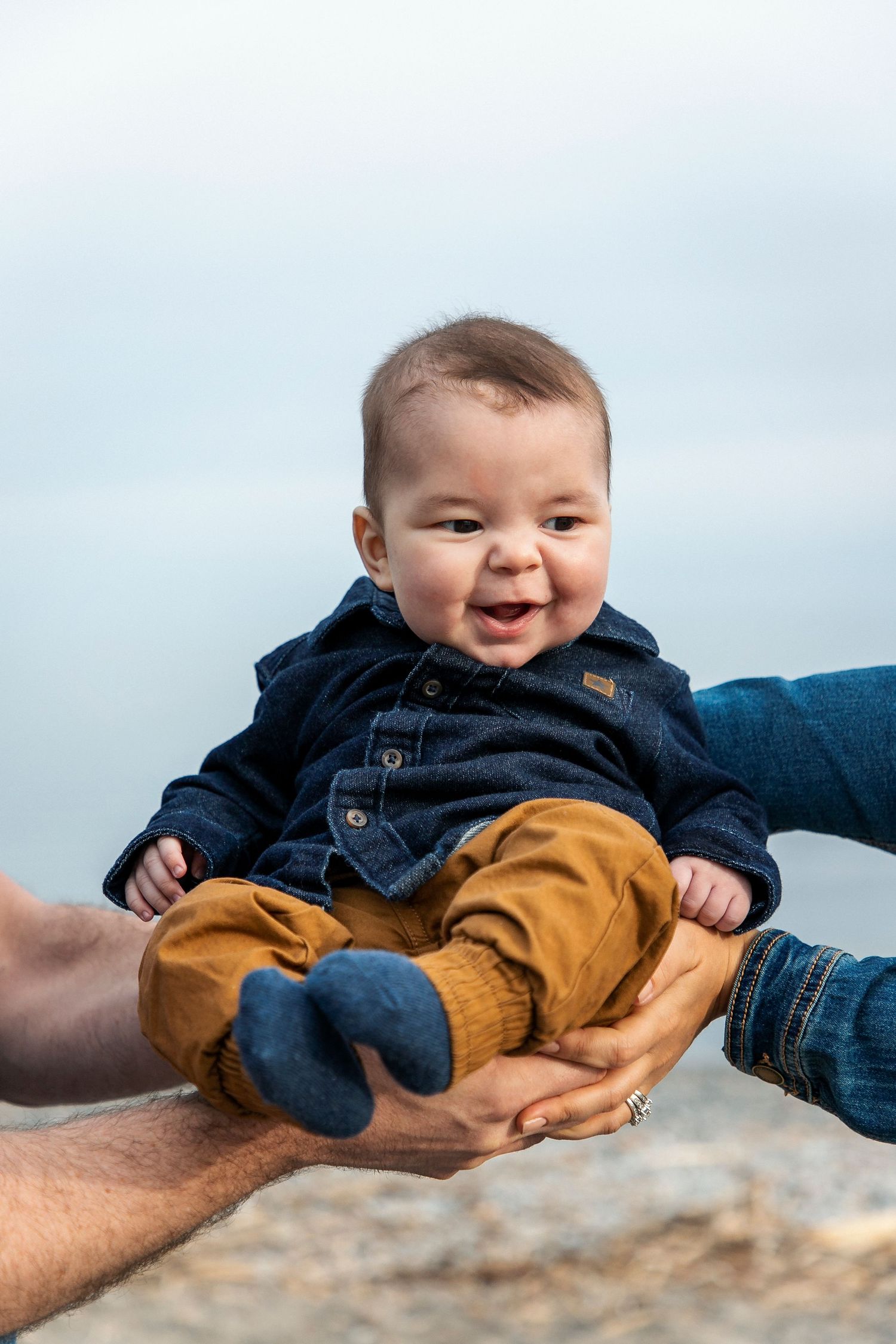 A baby with his mother and fathers hands holding him with the beach in the background