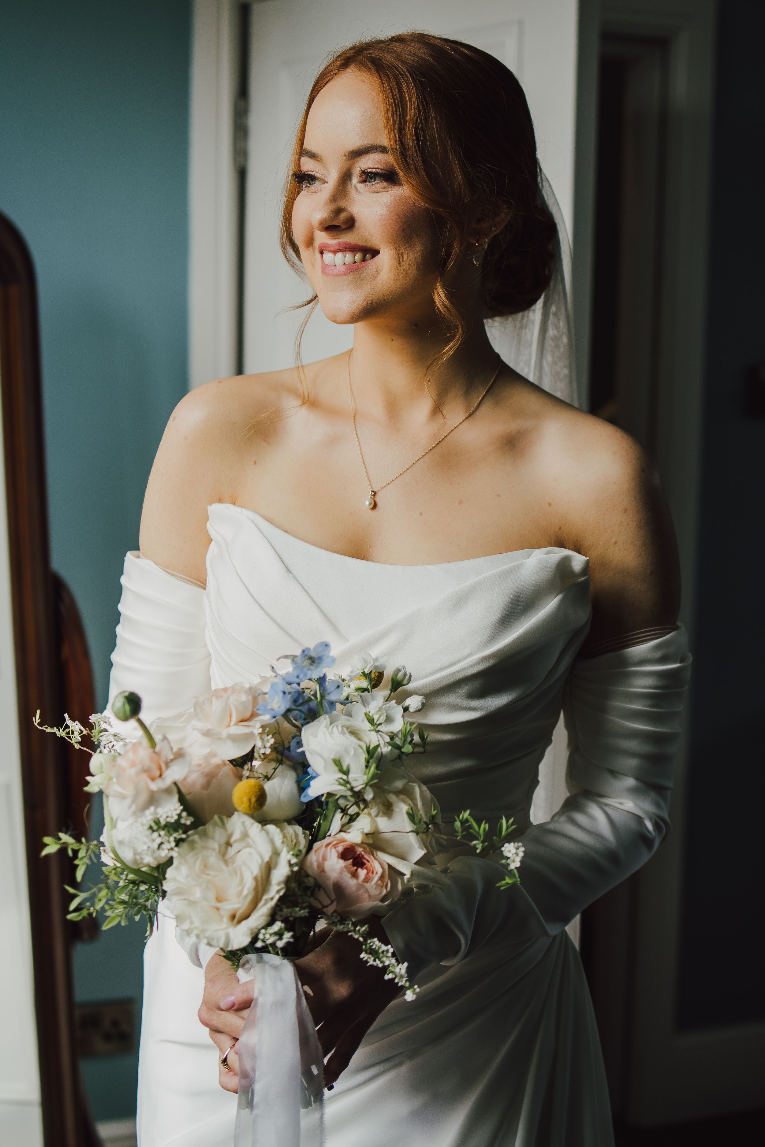 A bride in a white dress holds a beautiful bouquet of mixed flowers by a window.