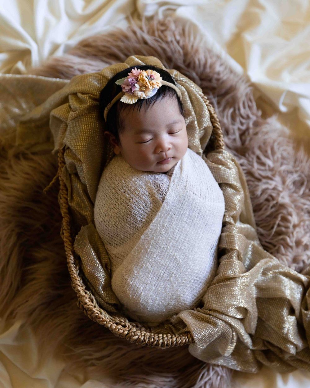 Sleeping newborn swaddled in white wrap with floral headband on textured beige backdrop.