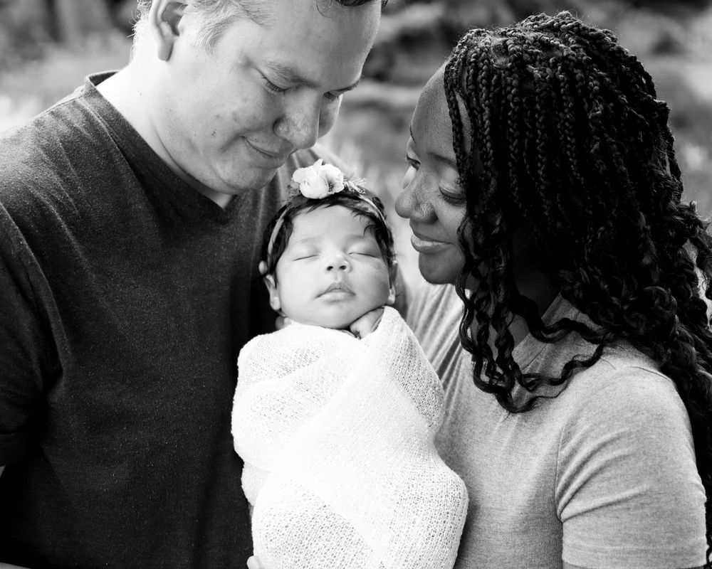 Black and white intimate family portrait of parents lovingly gazing at their newborn baby wrapped in white.