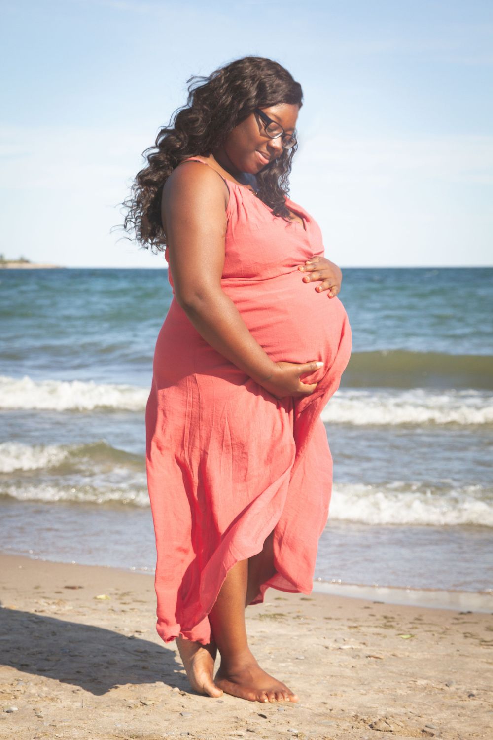 Expectant mother in coral dress standing on beach at sunset holding her baby bump.