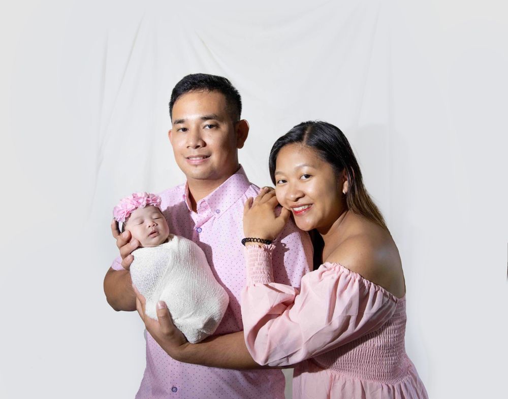 Family in matching pink outfits poses together with their newborn baby in a minimalist studio setting.