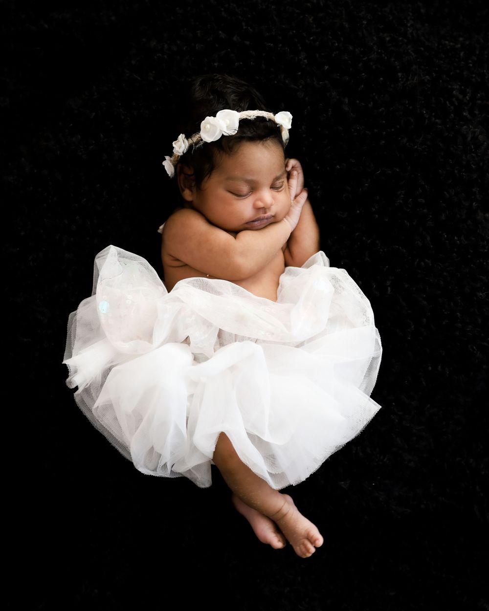 Sleeping baby in white tutu dress and flower crown posed on black background.