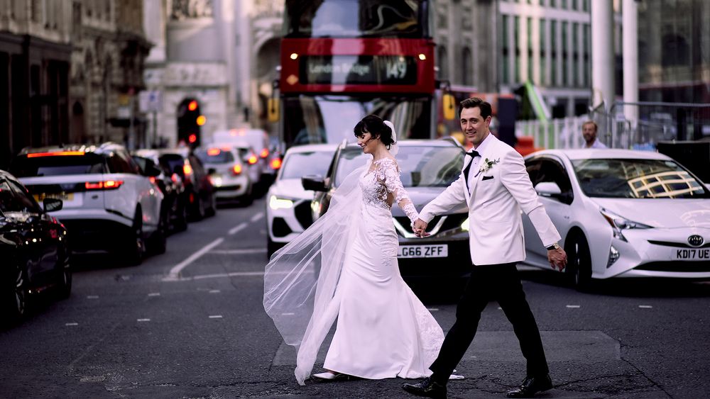 A couple in wedding attire playfully dances on a city street with a red double-decker bus in the background.
