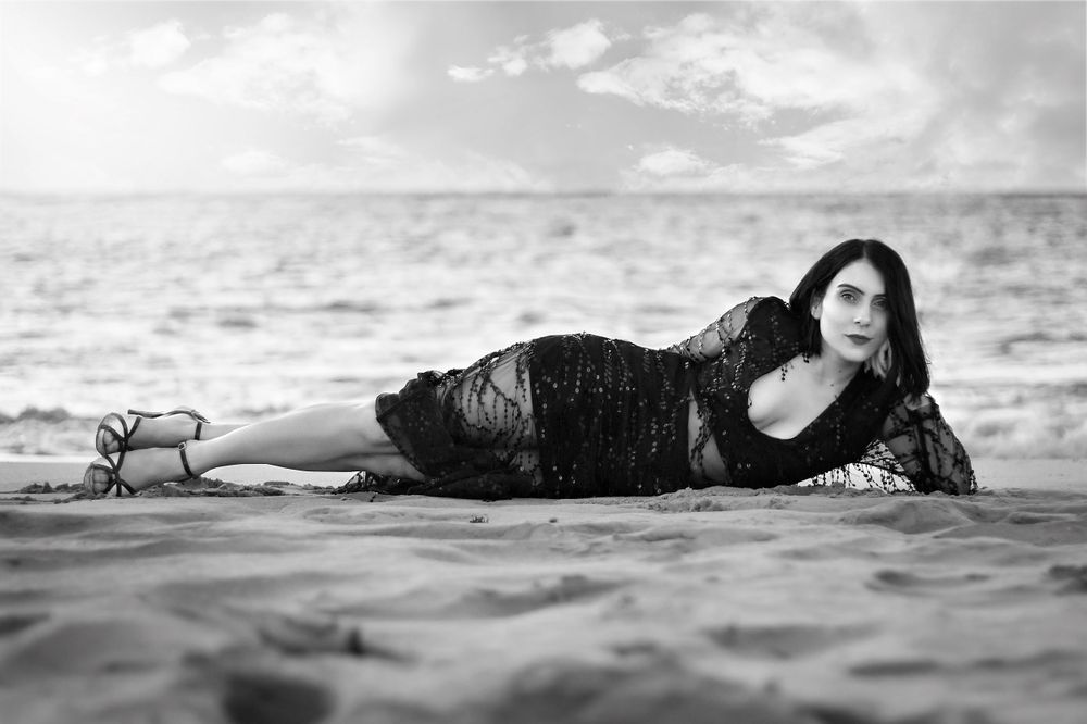 Black and White portrait of woman at beach in a beaded dress, laying on her side propping her head up with her hand.