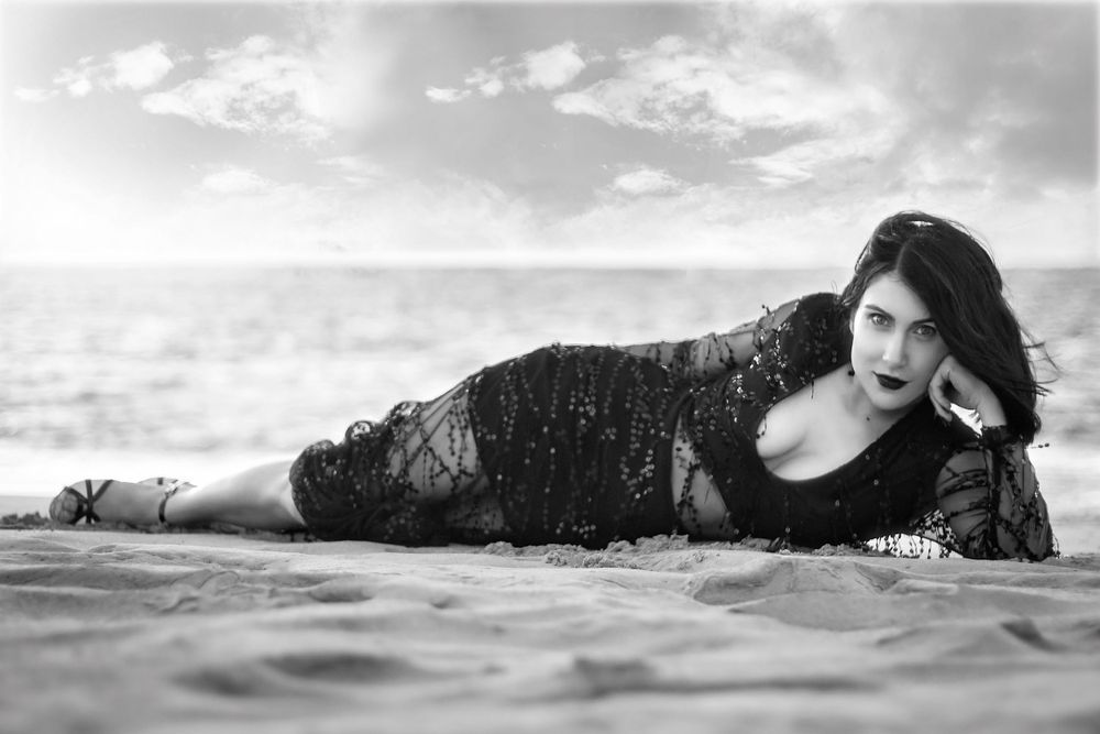 Black and White portrait of woman at beach in a beaded dress, laying on her side propping her head up with her hand.