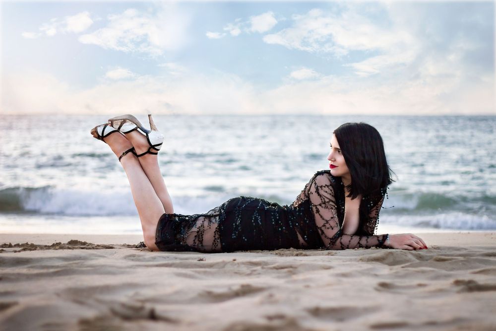 Dark haired woman in beaded evening gown, high heels, laying on her front on the beach with ocean in background.