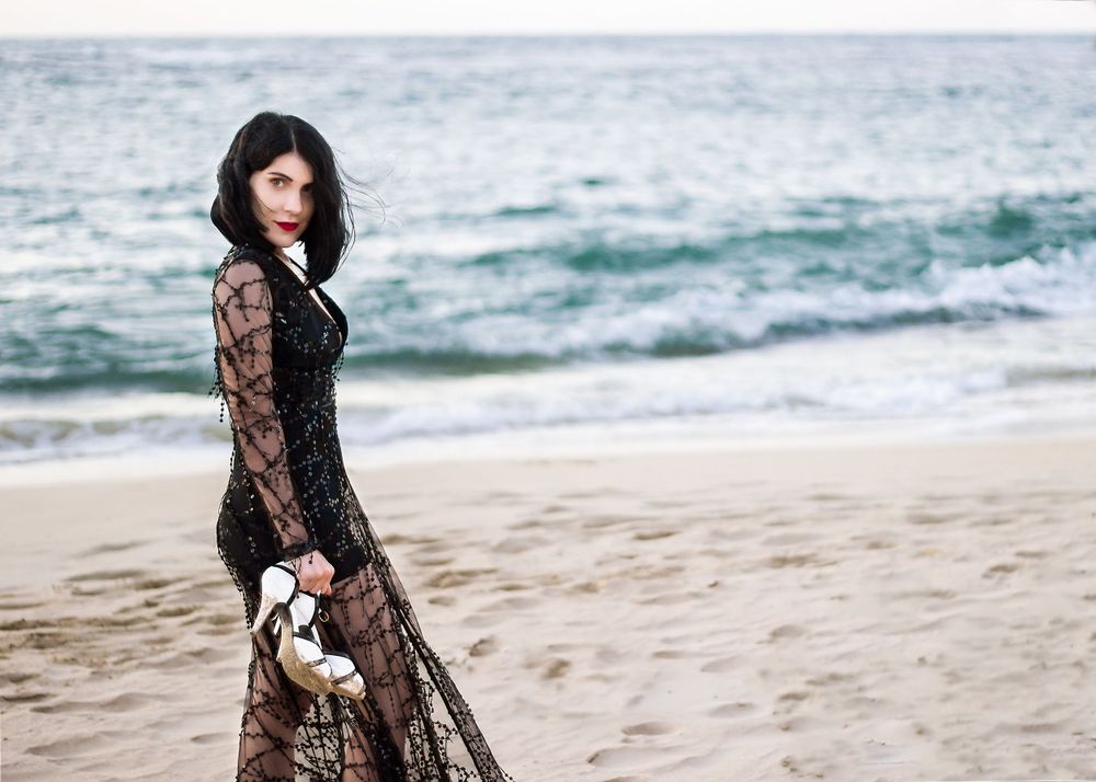 Beach landscape with a woman in beaded gown, carrying her black and white high heel shoes, off centre in foreground.