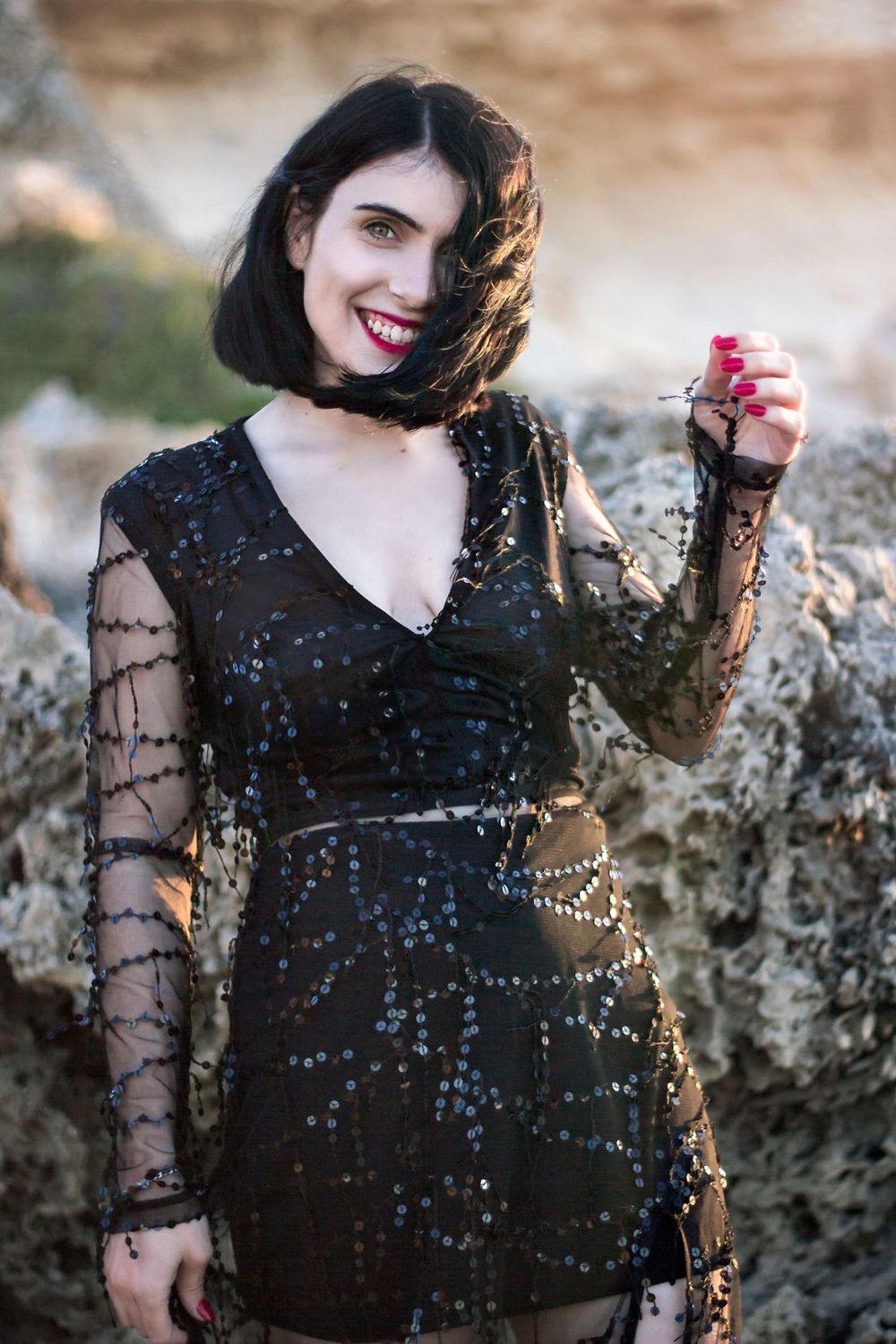 Dark short haired woman wearing bright red lipstick and beaded evening gown smiling at viewer while wind blows her hair.