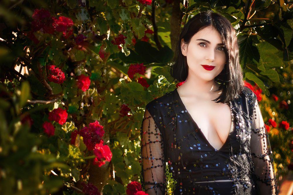 Woman with short black hair wearing bright red lipstick and black beaded gown standing in front of wall red rose bushes.
