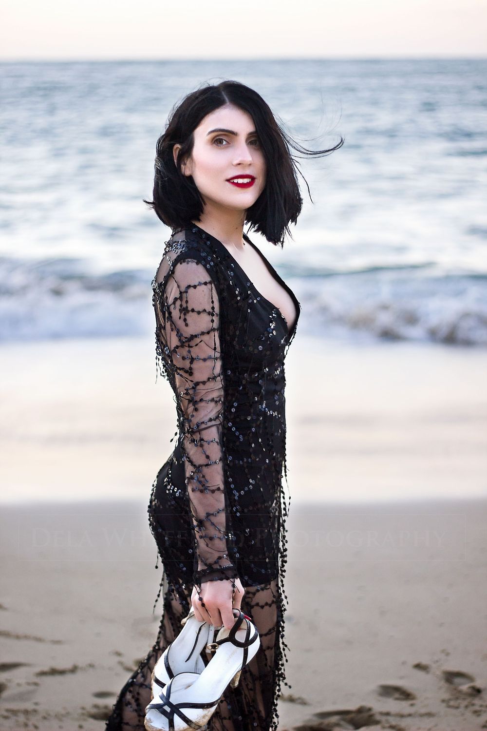 Woman in beaded gown at sunset on beach, carrying her high heel shoes with the ocean in the background.