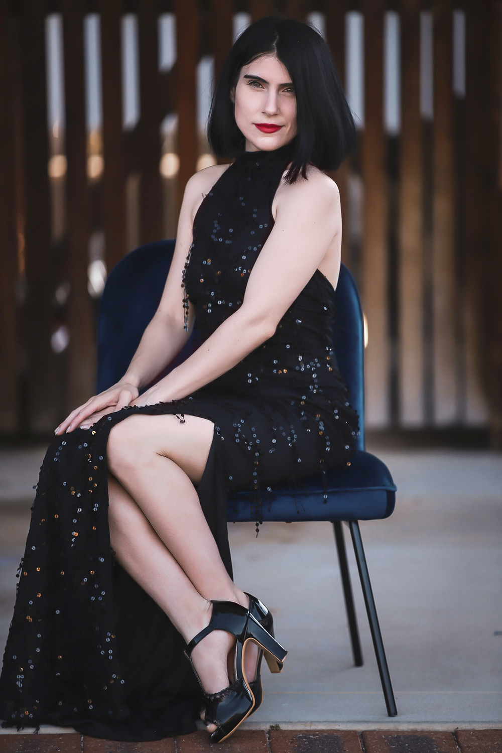 Woman wearing bright red lipstick and black evening gown with high slit on leg, sitting modestly on blue velvet chair.