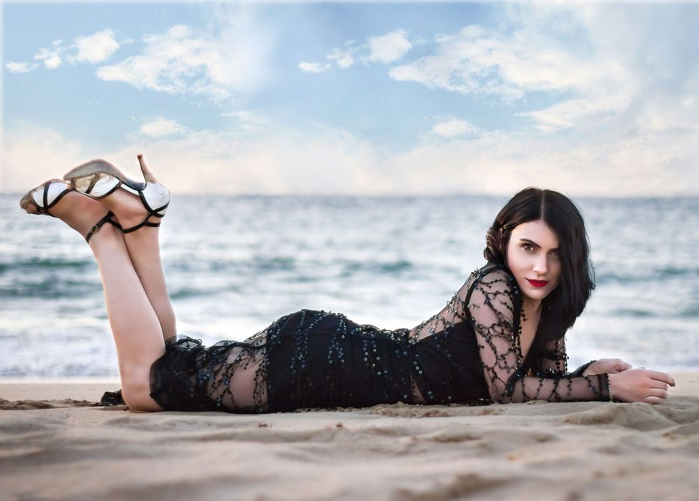 Dark haired woman in beaded evening gown, high heels, laying on her front on the beach with ocean in background.