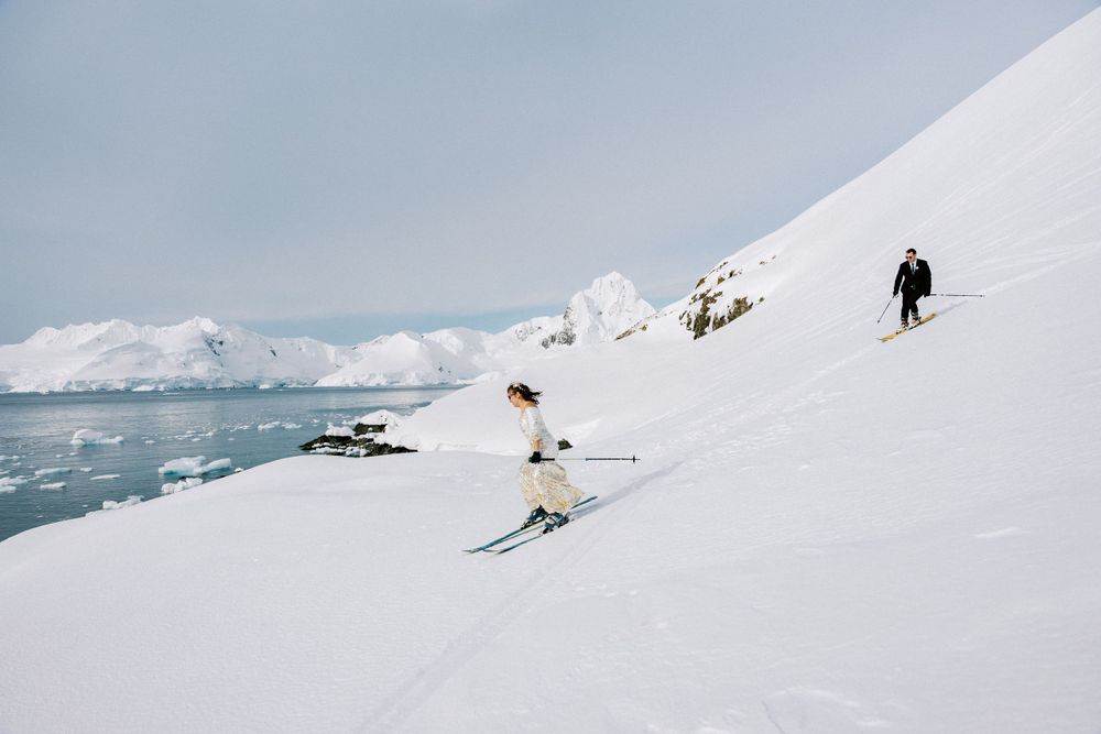Skiers descend a snow-covered slope alongside icy Antarctic waters.