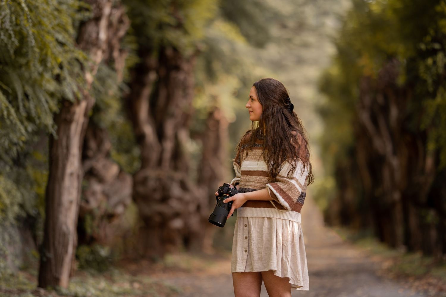 Hounds & Horizons photographer Laura Holzner holding a camera smiling on a tree-lined dirt road in natural light.