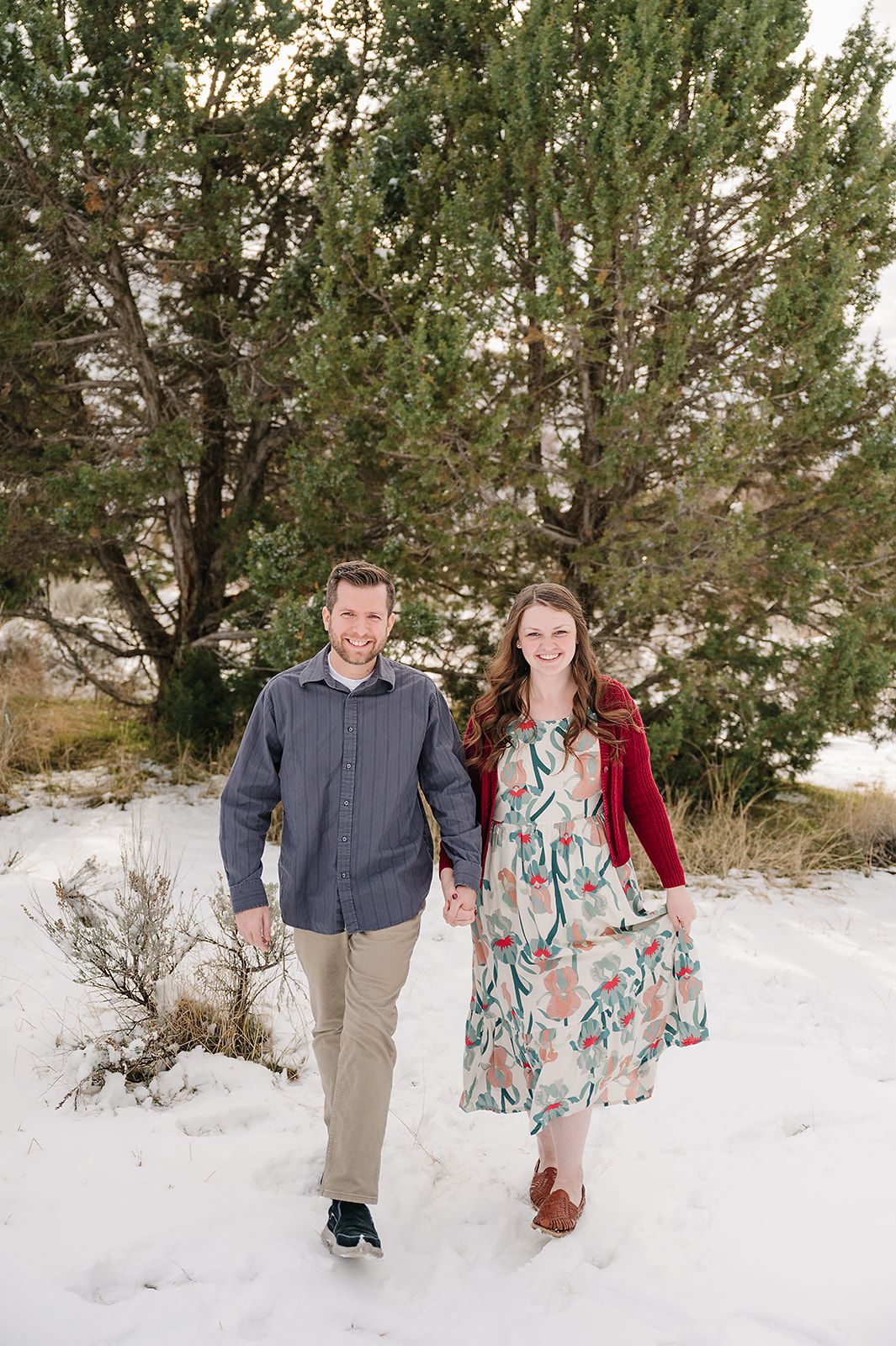 A couple walks hand in hand through snow-covered terrain with evergreen trees in the background.