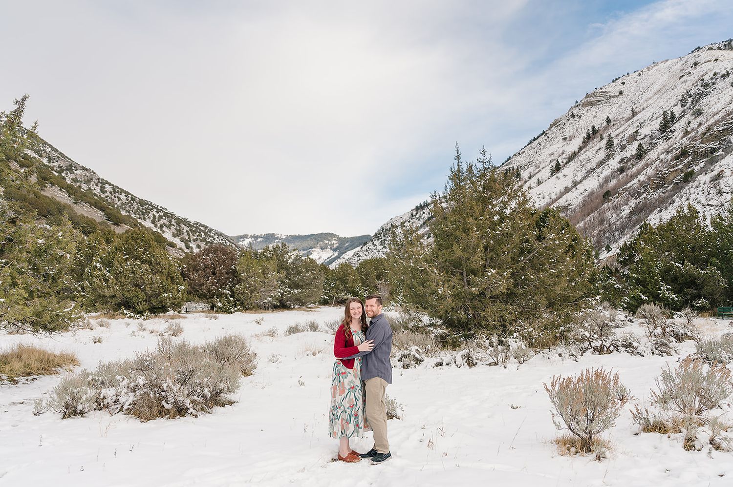 Two people stand together on a snowy mountain trail surrounded by evergreen trees and rocky peaks.