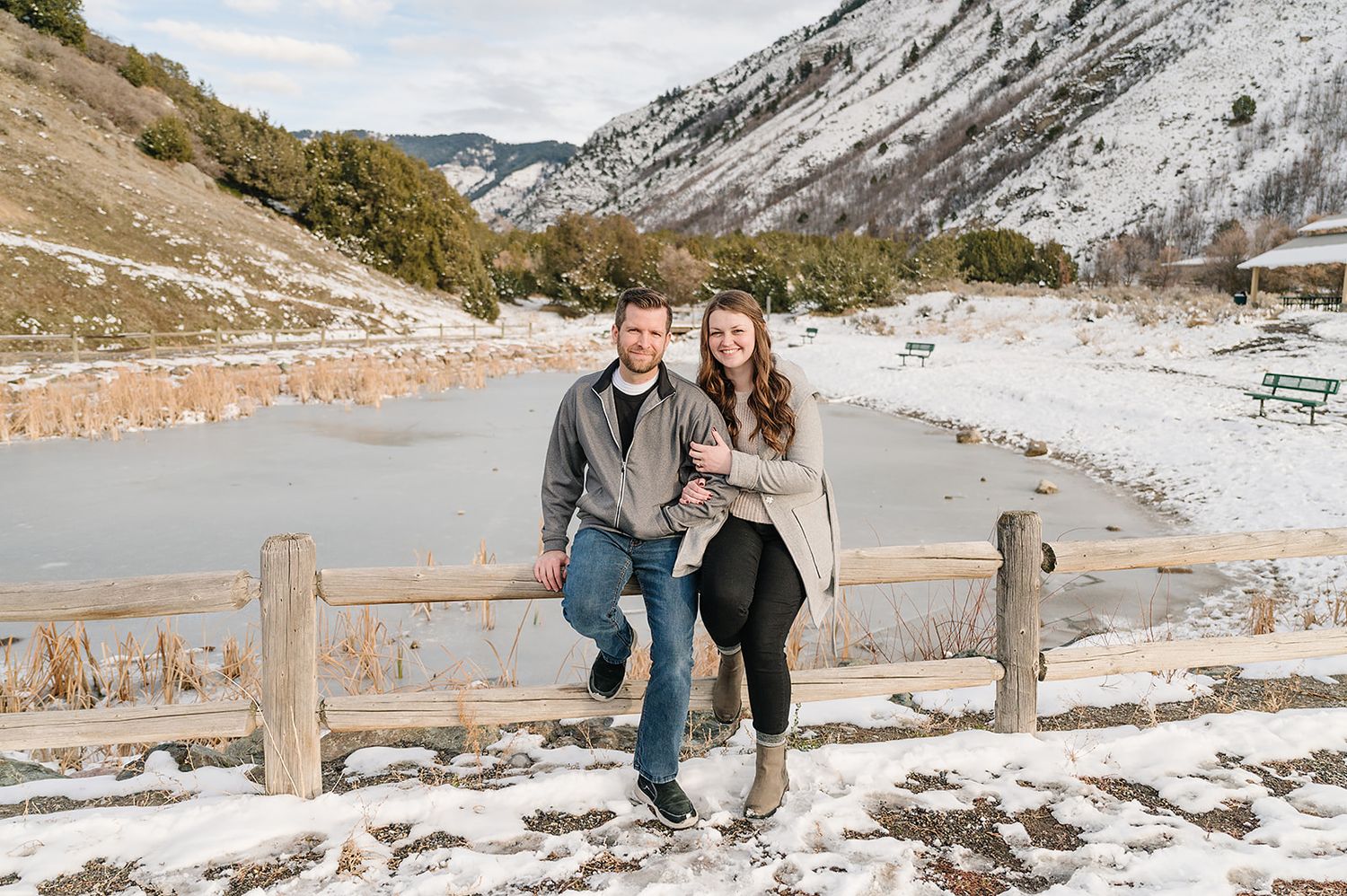 A couple poses together on a wooden fence with snow-covered mountains in the background on a winter day.