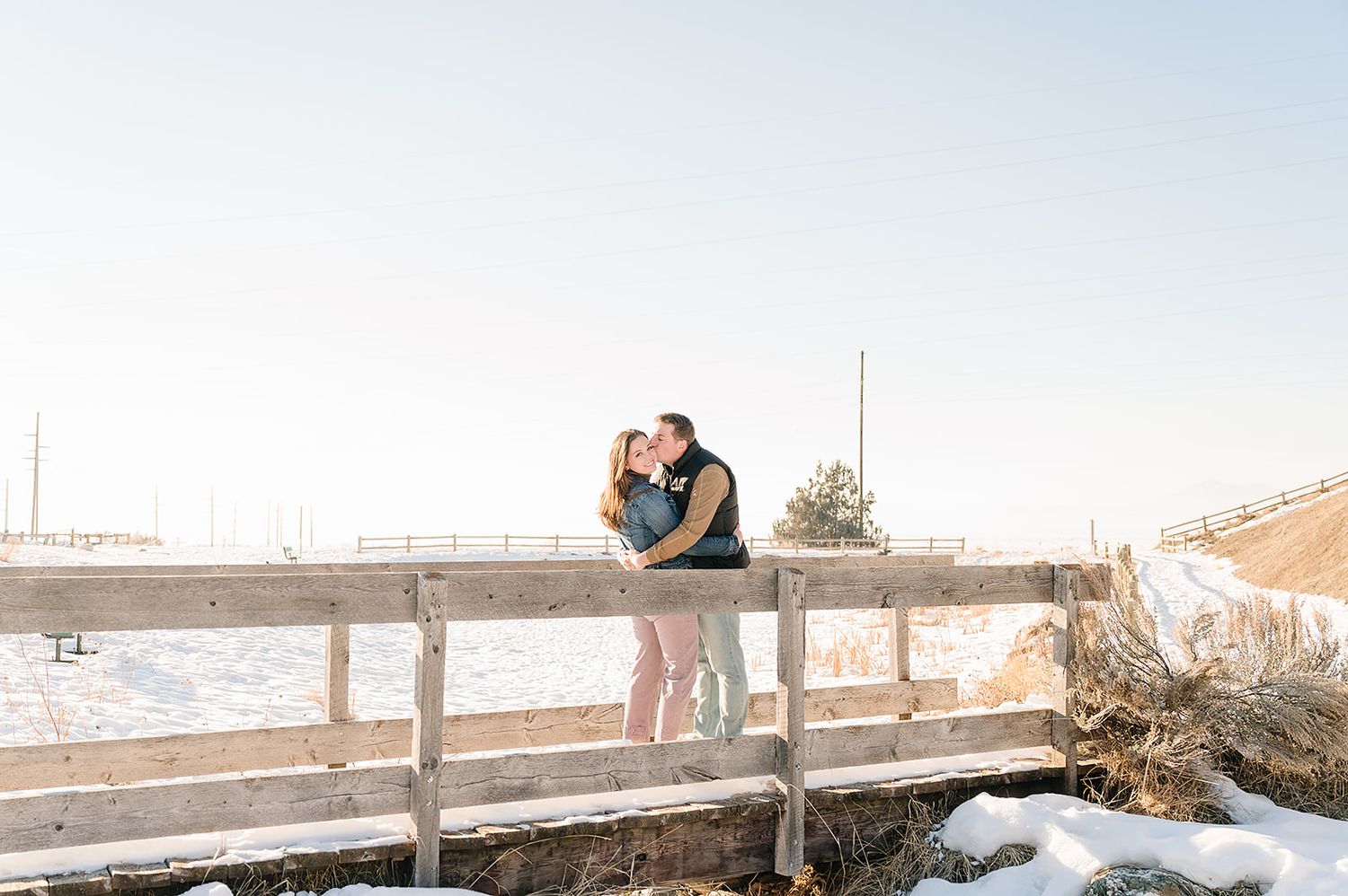 Couple standing on wooden bridge surrounded by winter snow and mountains.