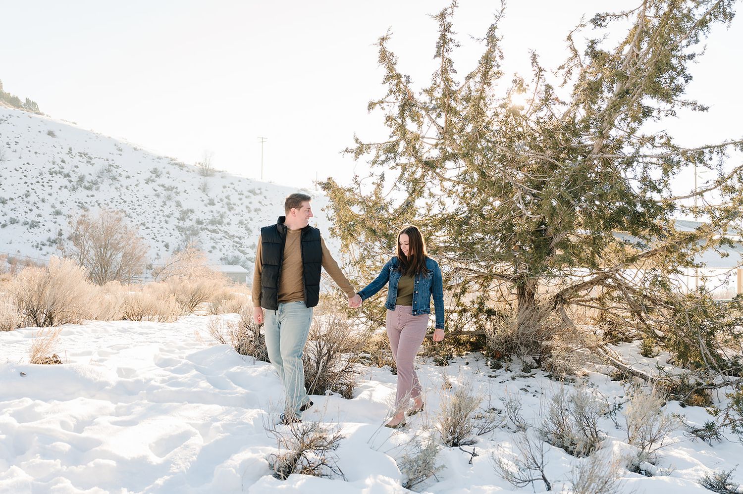 Two people holding hands while walking through a snowy mountain landscape at sunset.