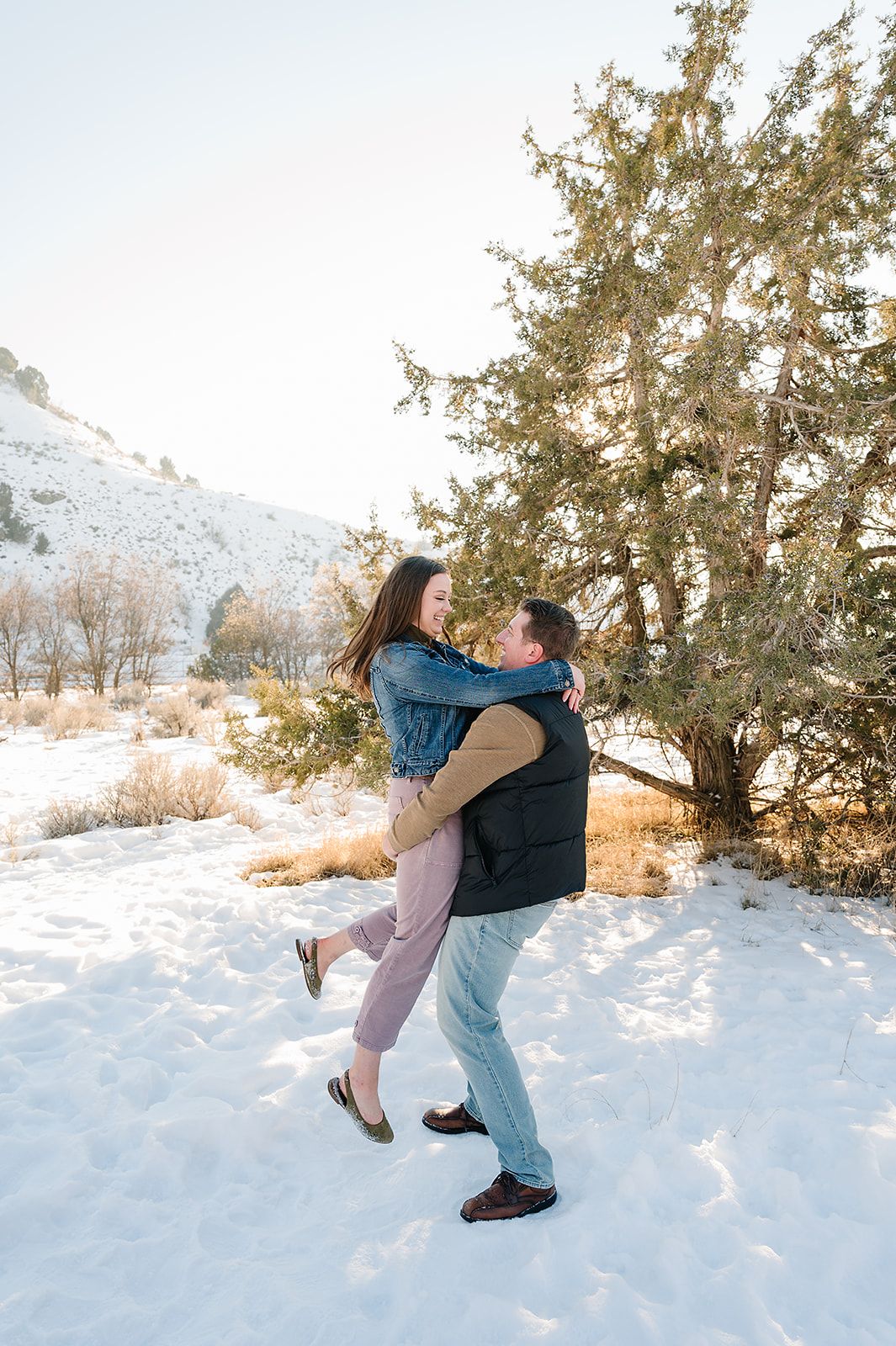 Playful couple embrace in a snowy winter landscape with evergreen trees.