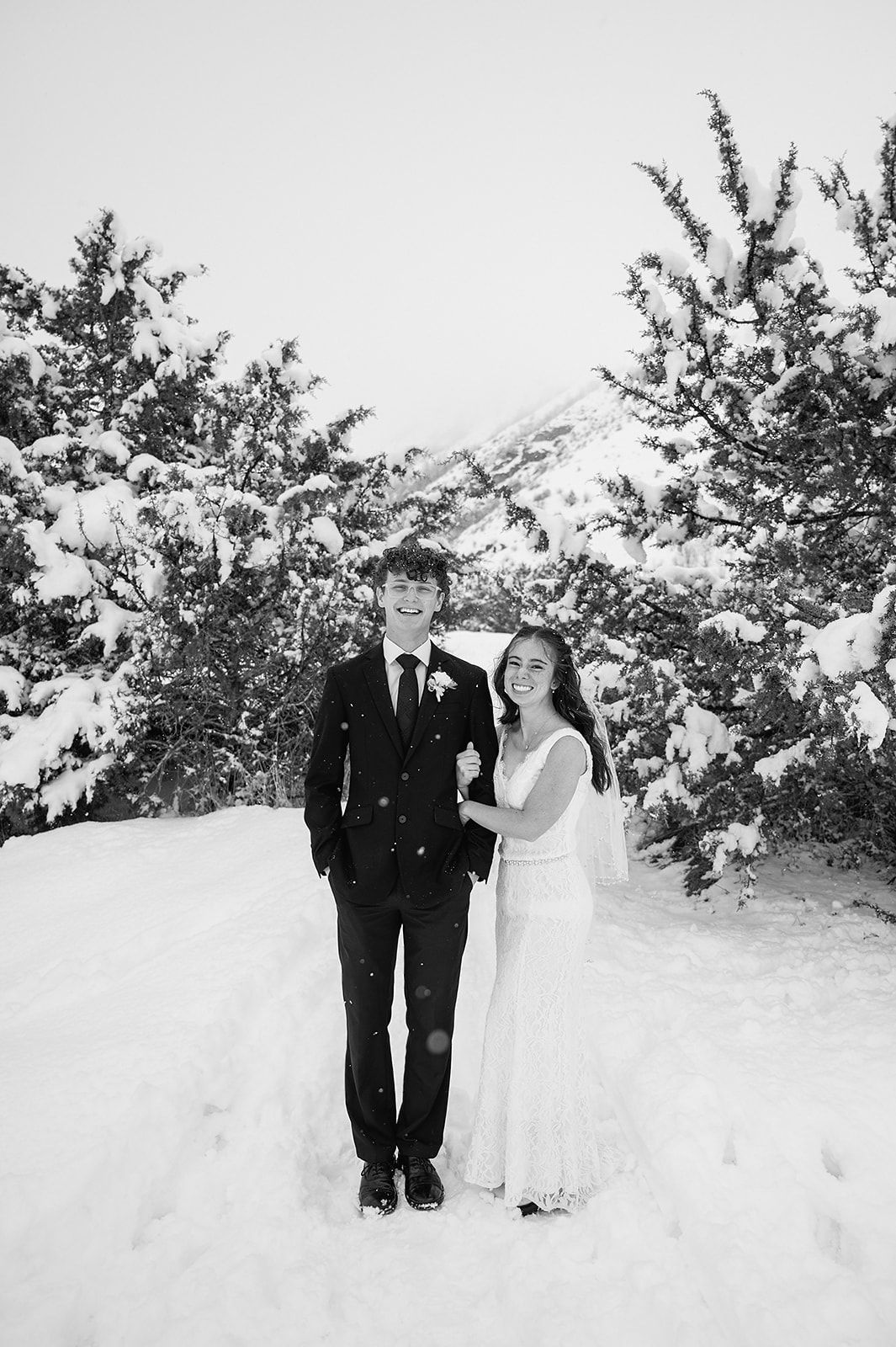 Wedding couple poses in snow-covered mountain setting with evergreen trees.