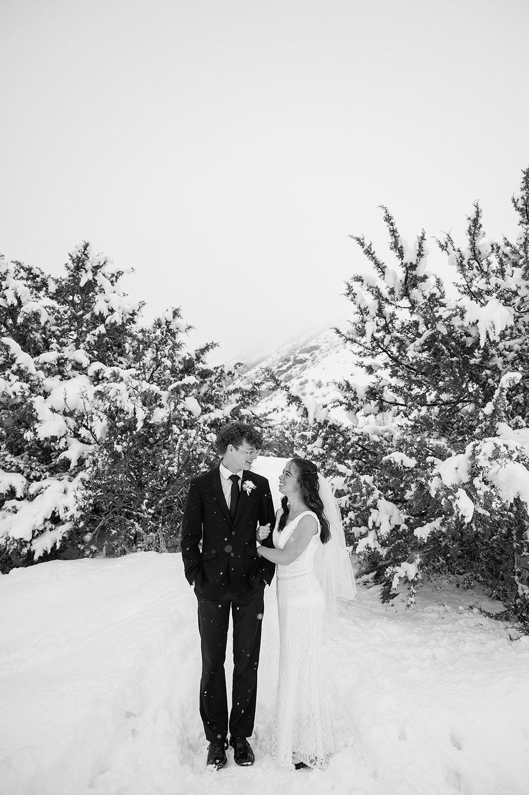 Black and white photo of newlyweds standing in snow-covered landscape with evergreen trees.