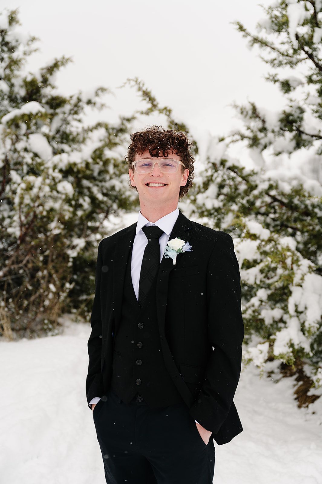 Person in black suit with boutonniere stands in snowy winter setting.