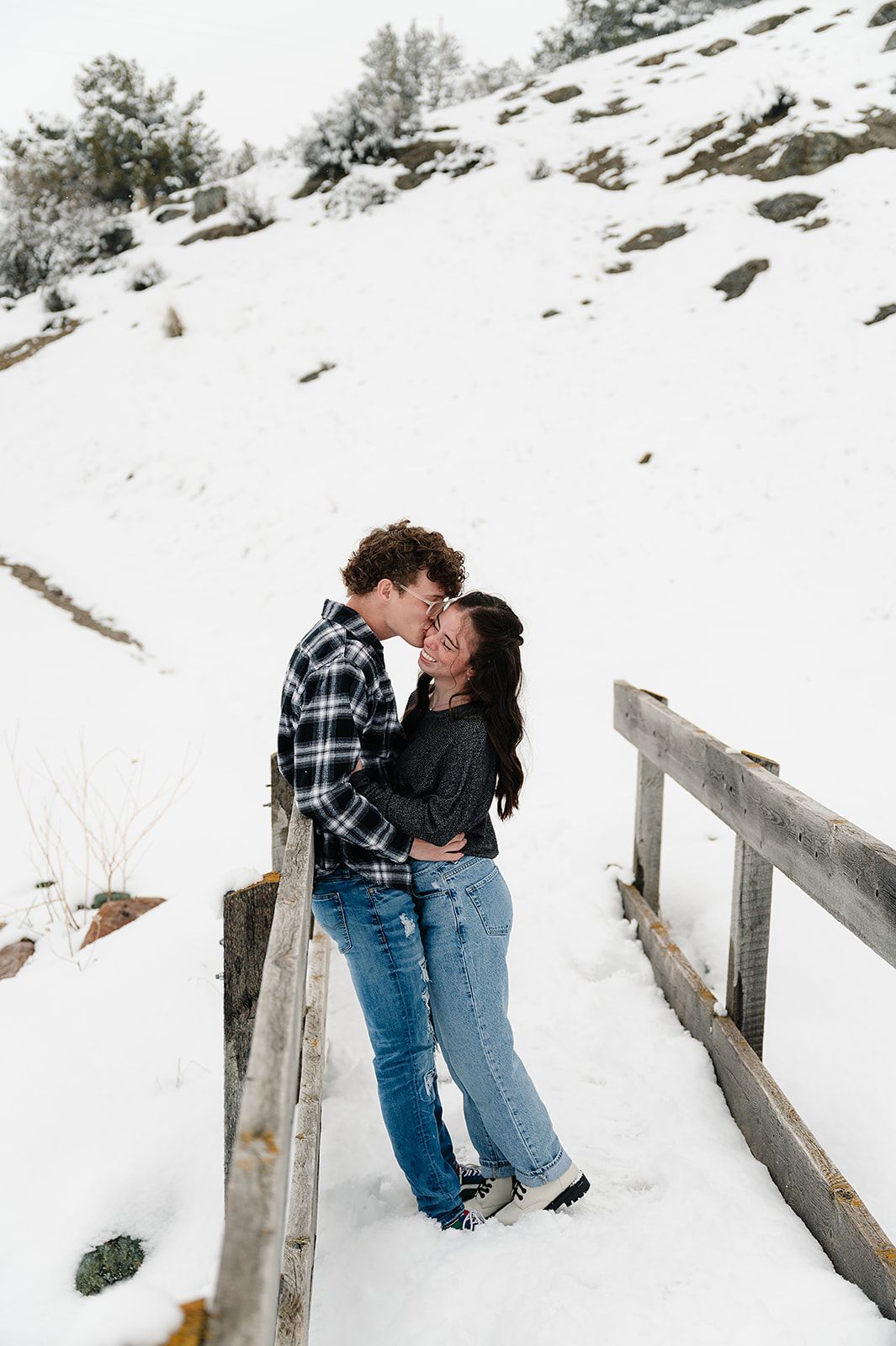 Couple sharing romantic moment on snowy bridge with mountain backdrop.