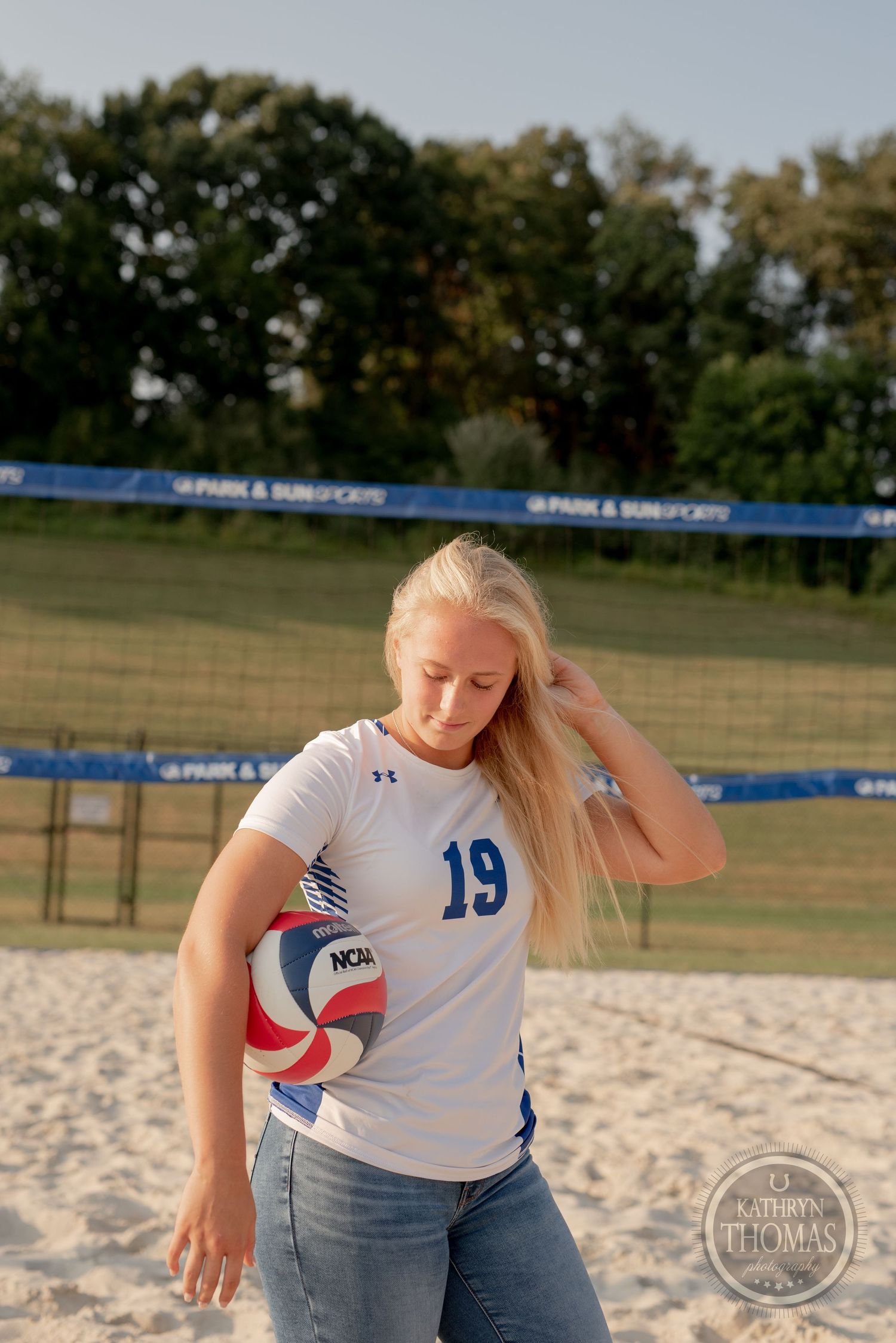 An athlete in jersey number 19 practices with a volleyball on a sandy outdoor court.