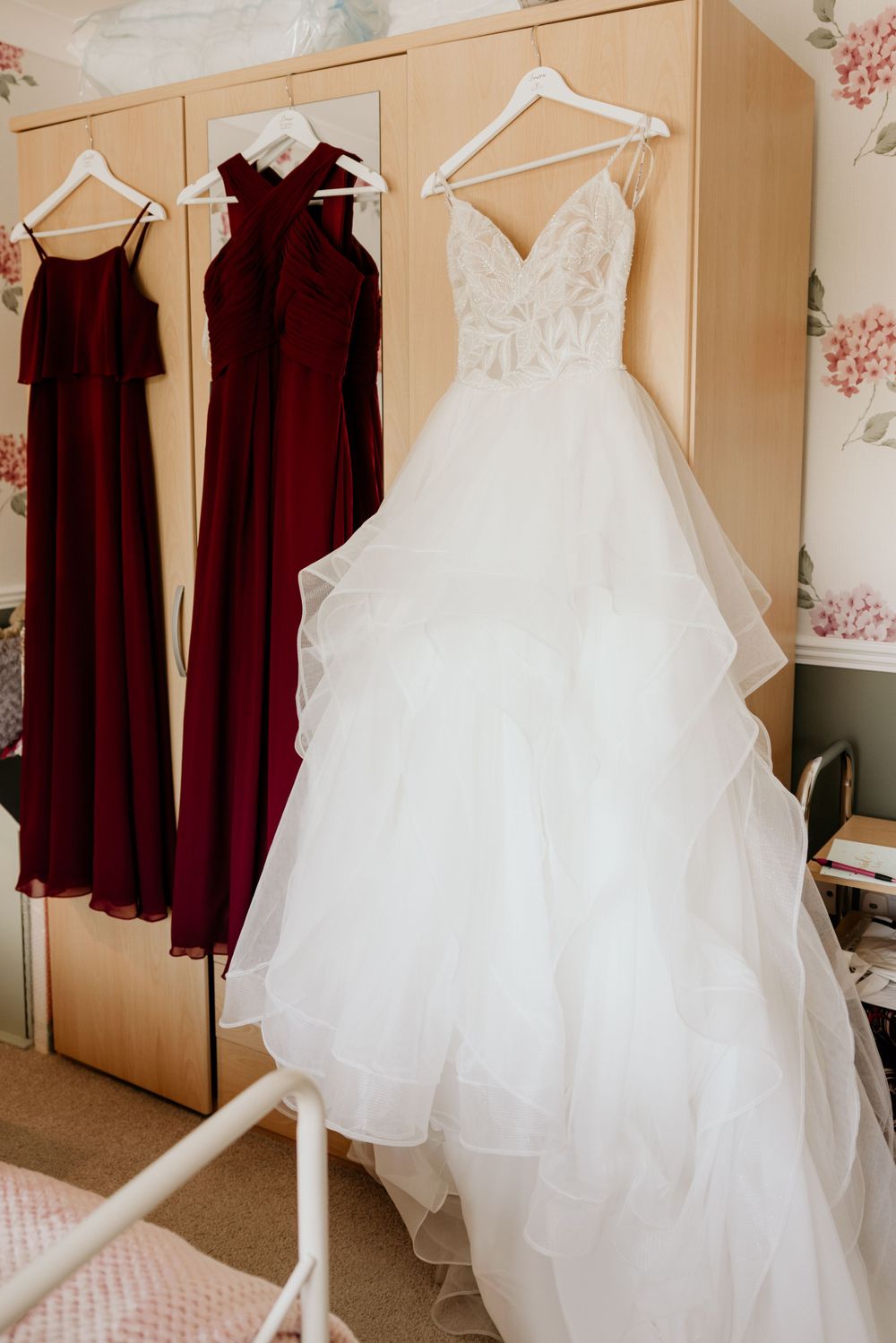 Wedding dress and burgundy bridesmaid dresses hanging on wooden hangers against floral wallpaper.