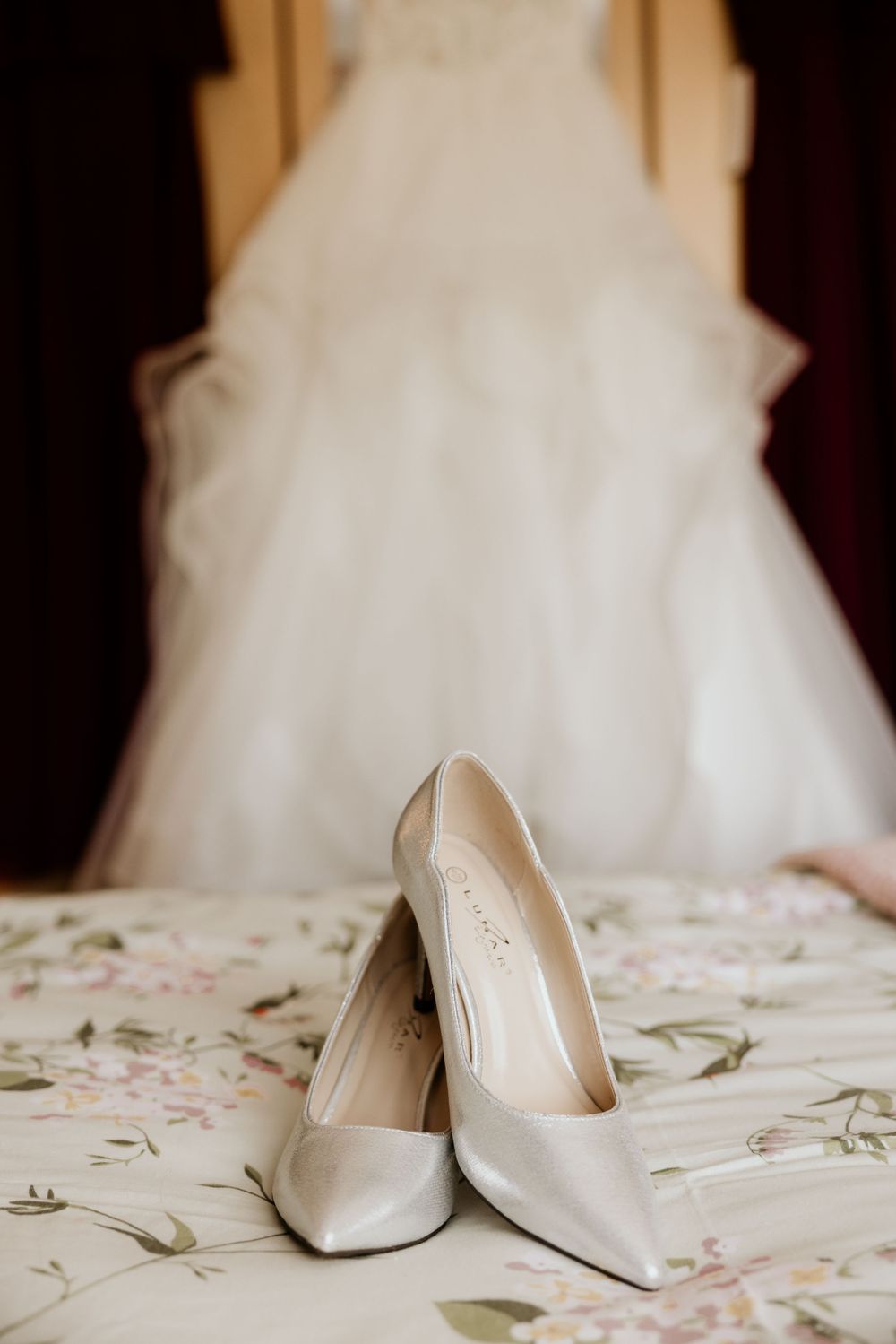 Elegant white bridal heels placed in front of flowing wedding dress on floral print surface.