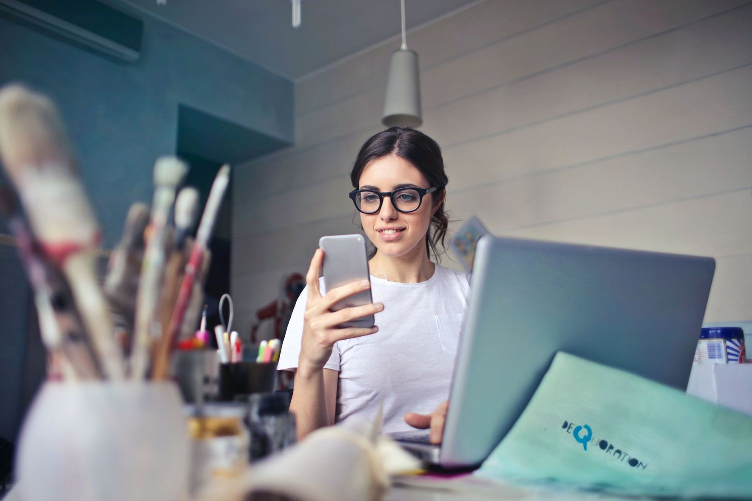 Professional looking at smartphone while working at desk with laptop and office supplies.
