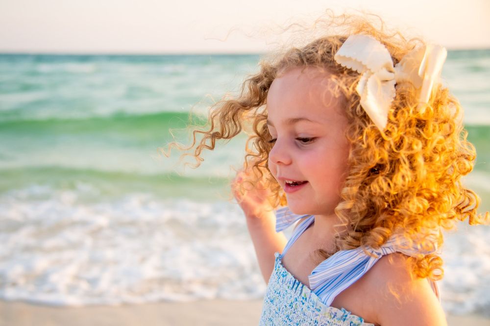 Young child with curly blonde hair and a bow enjoying the beach on a sunny day with turquoise waves in the background.