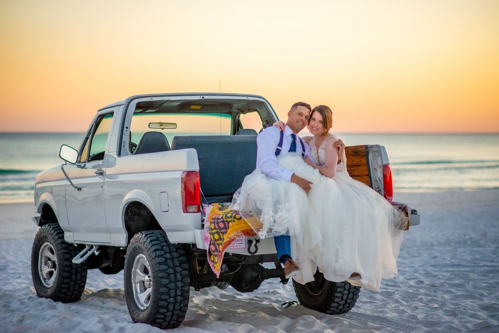 Newlyweds sit on tailgate of lifted white truck at beach during sunset.