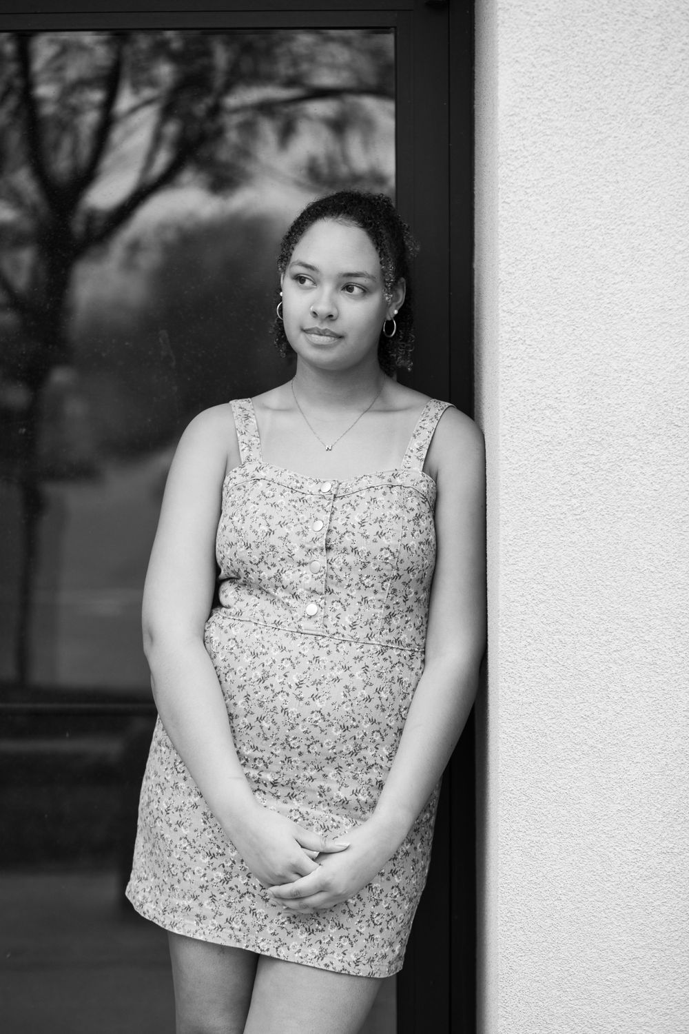 A black and white portrait of someone in a floral dress standing by a window.