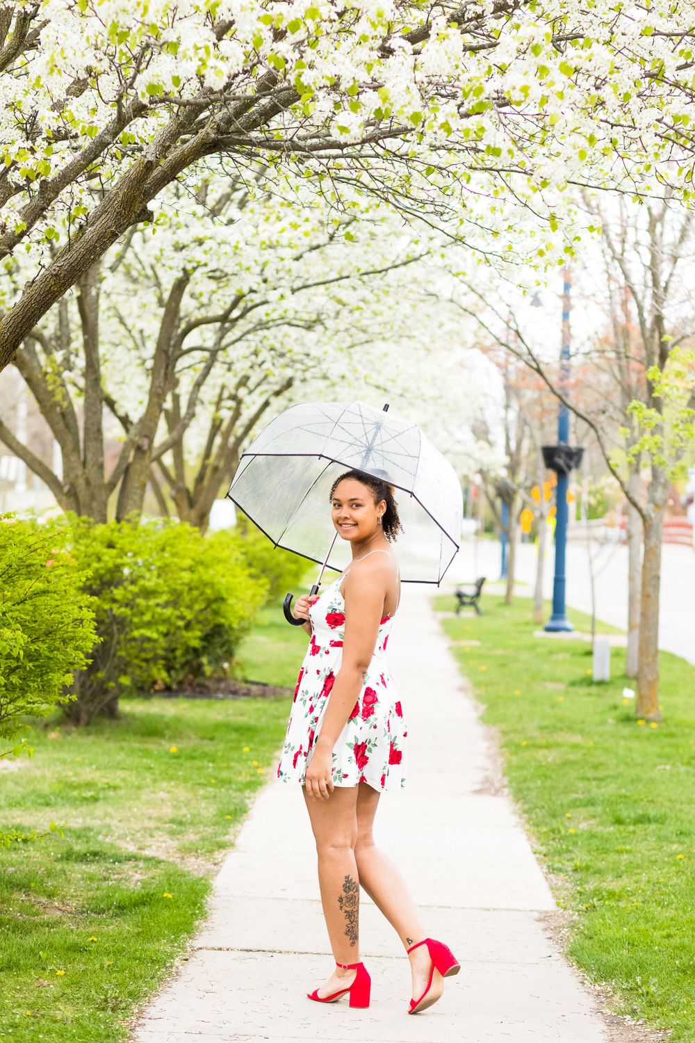 Someone in a floral dress and red heels twirls with a white parasol on a spring sidewalk.