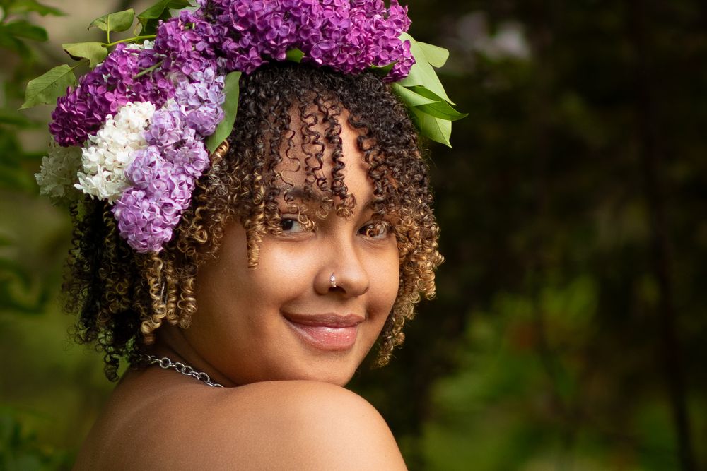 A natural portrait with purple lilac flowers adorning curly hair in a garden setting.
