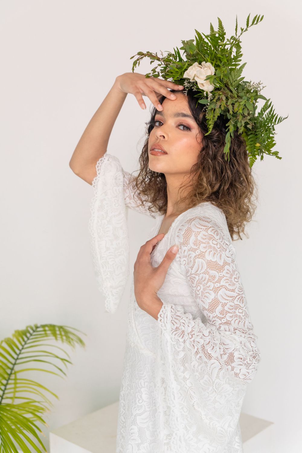 A model in a white lace dress poses with a floral crown and greenery in a minimalist studio setting.