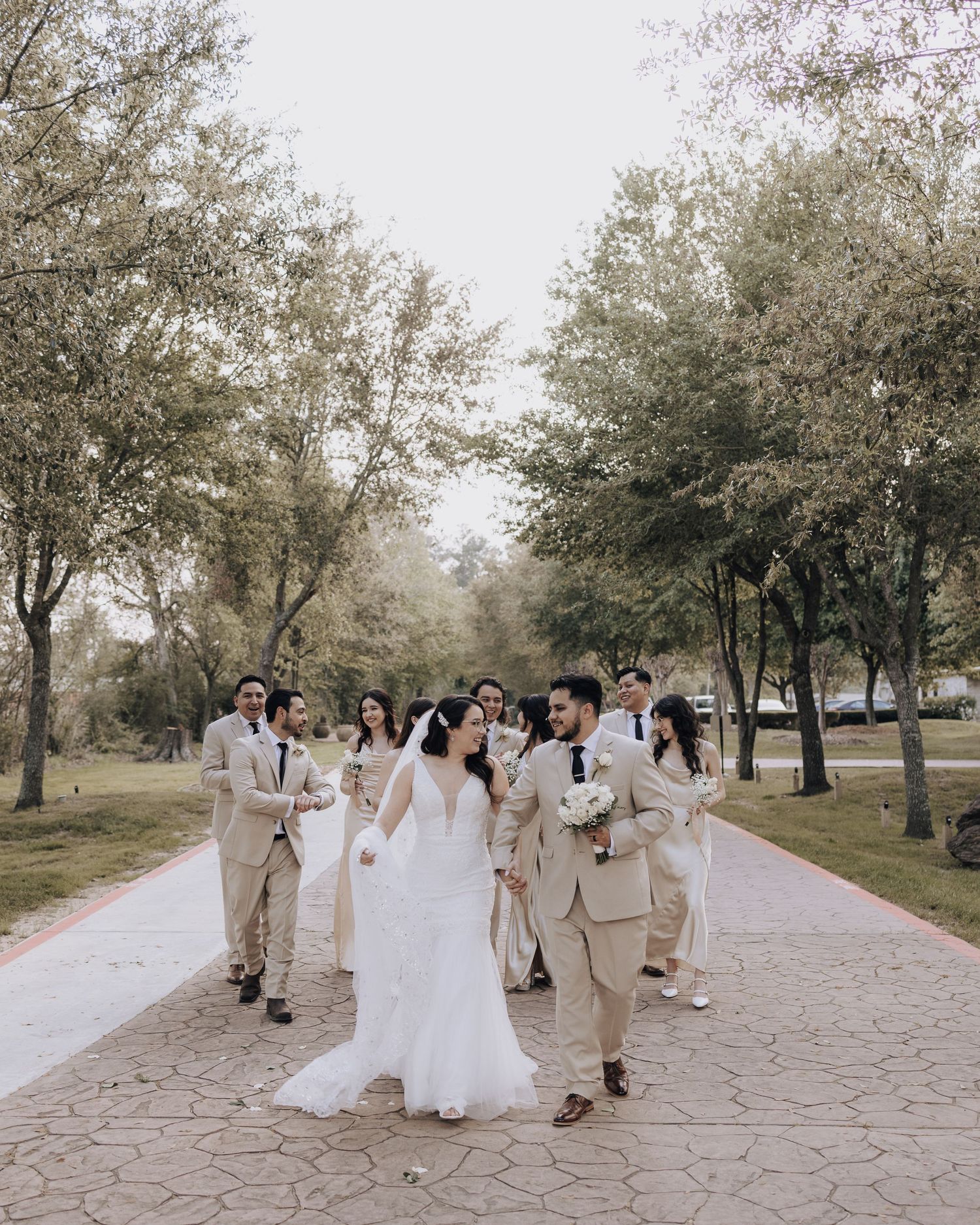 Bridal party strolls down outdoor pathway surrounded by trees in matching neutral colors.