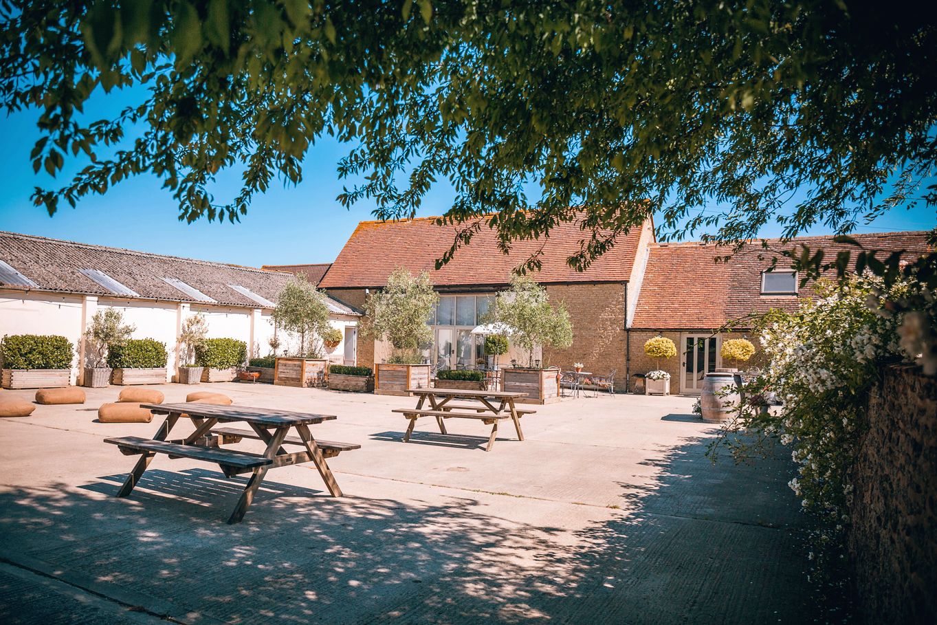 Sunny courtyard of a rustic venue with wooden picnic tables and traditional farm buildings.