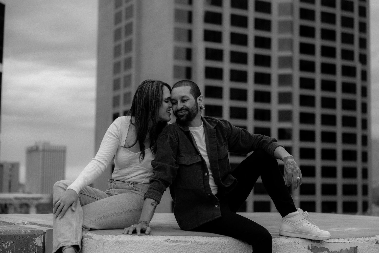 A black and white photo of a couple sitting close together on a rooftop with city buildings behind.