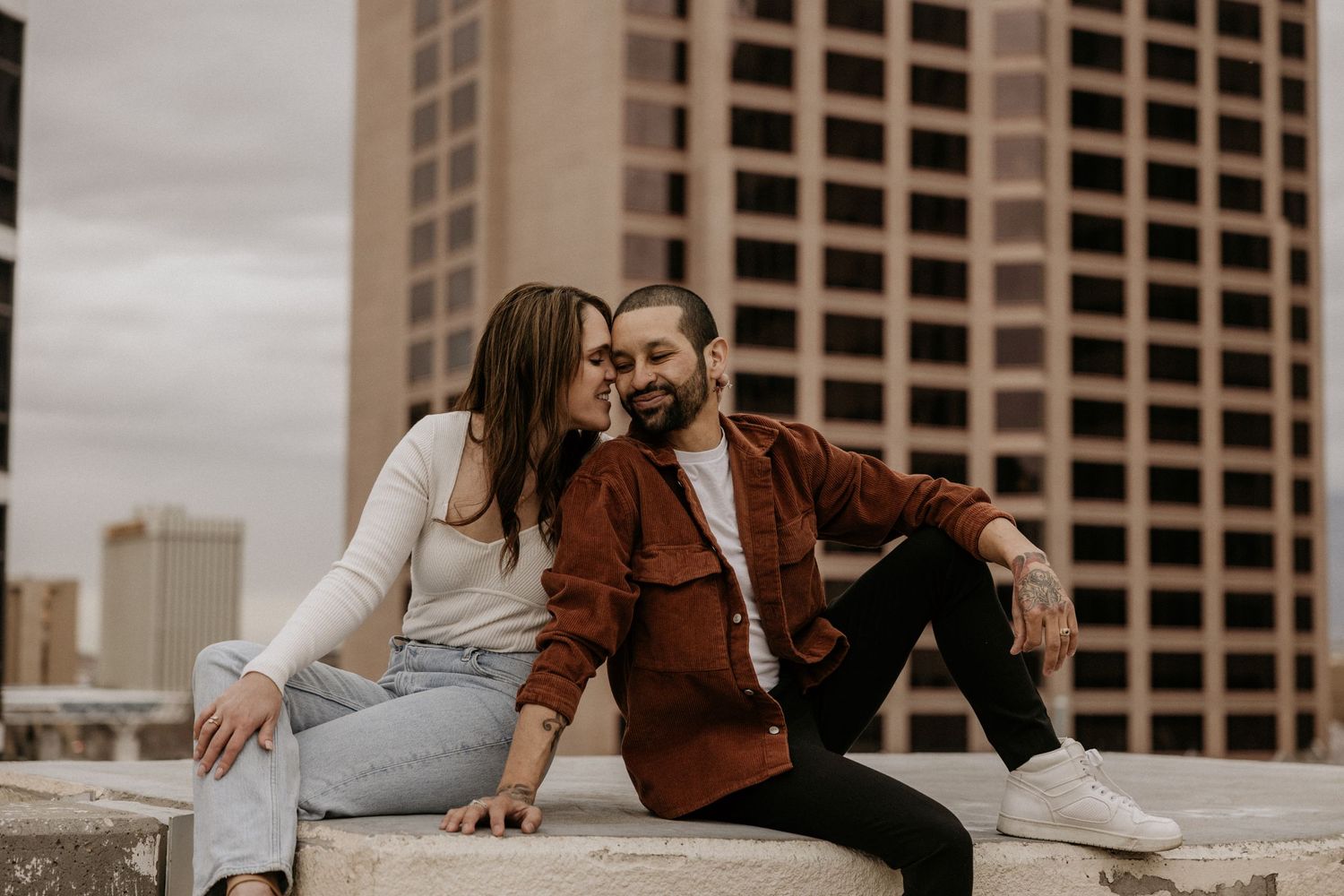 A couple sits together on a rooftop ledge, smiling and leaning close with city buildings in the background.