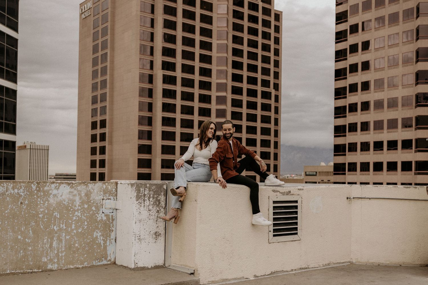 Romantic couple sitting together on rooftop wall with tall city buildings in background.