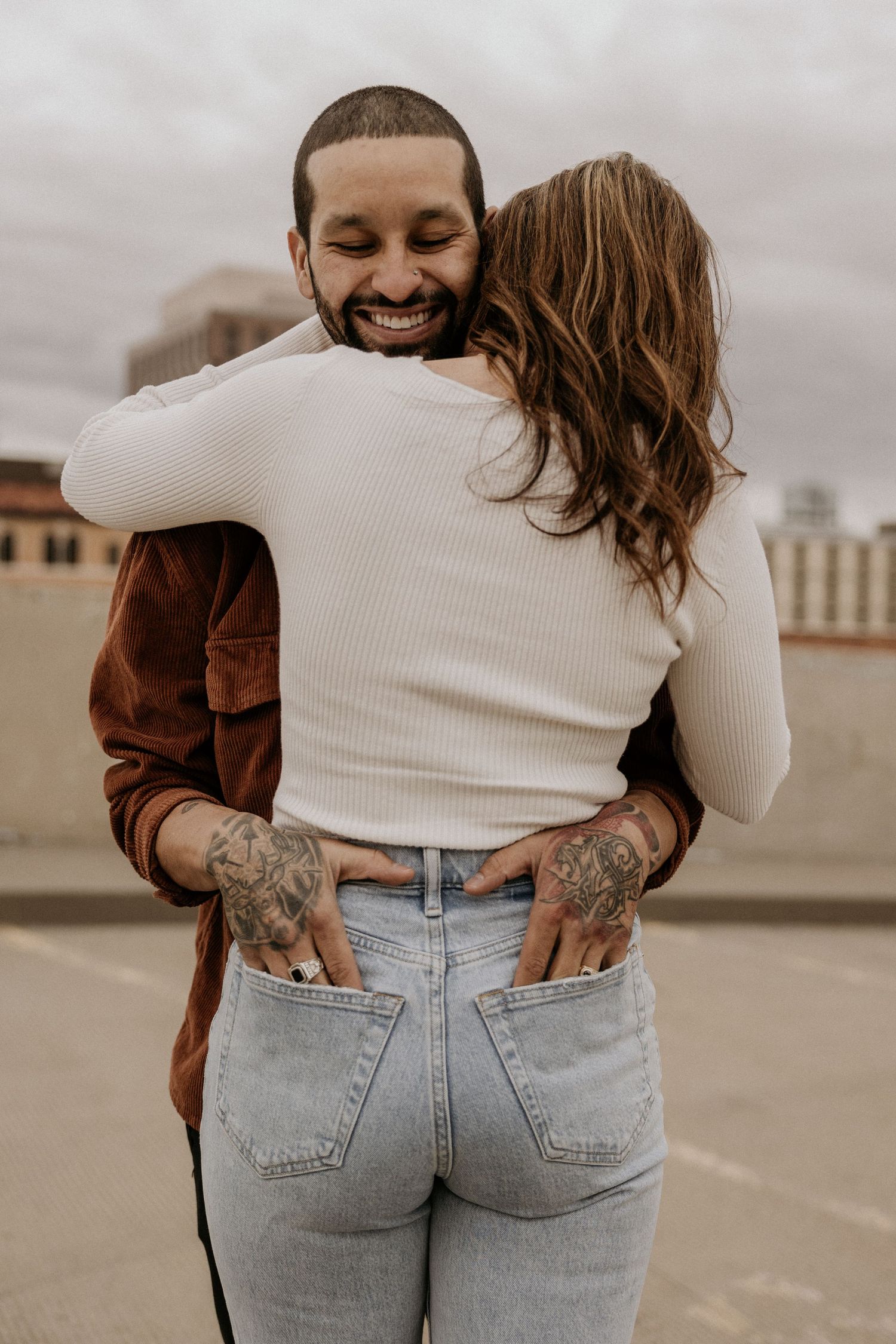 Smiling tattooed man hugging woman in white ribbed top on urban rooftop.