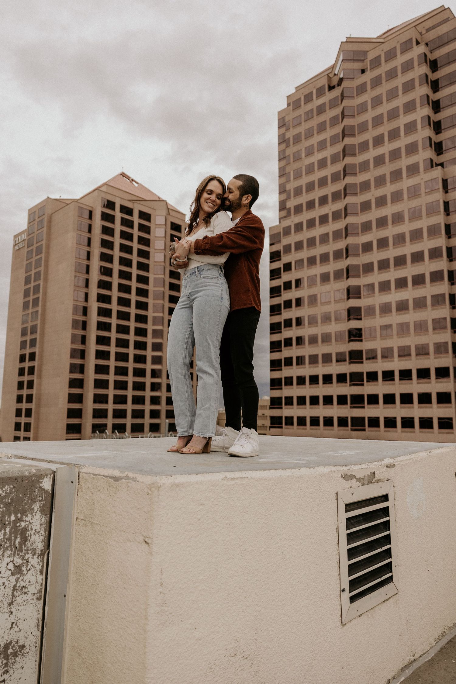 Couple embracing and standing on rooftop with large urban skyscrapers under overcast sky.